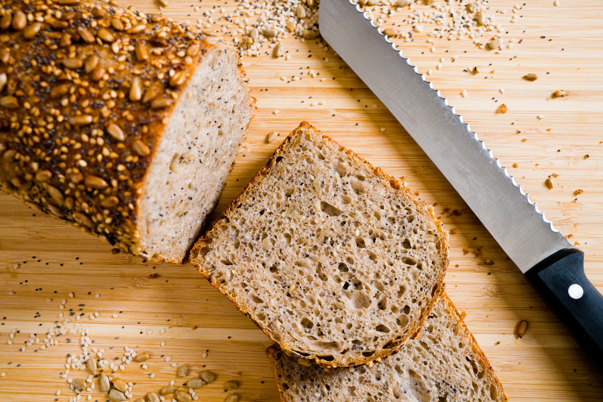 Two slices of seeded sandwich bread sit on a wooden cutting board with the remaining whole loaf to the left and a bread knife to the right.