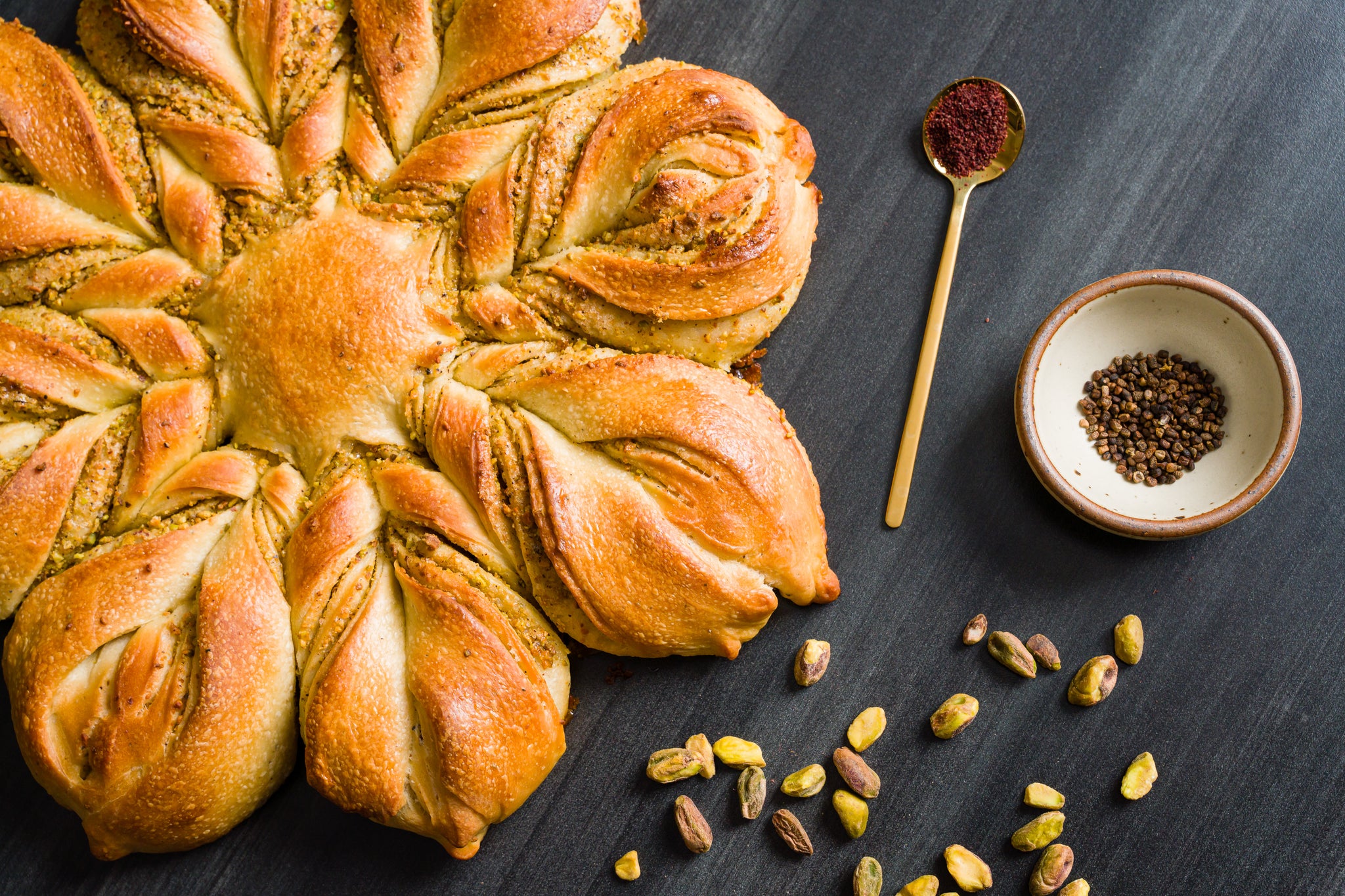 Pistachio cardamom star shaped bread on black countertop. To the right is a scattering of pistachios, a small white bowl of cardamom, and a gold teaspoon containing sumac