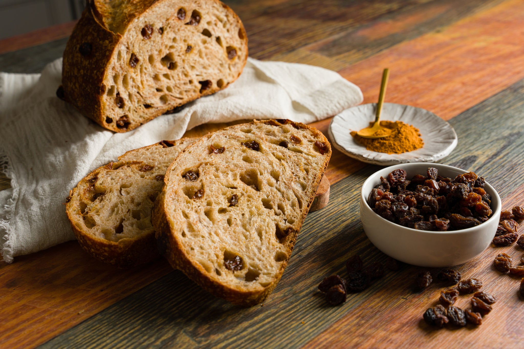 Slices of cinnamon raisin sourdough on a wooden table. A small dish of cinnamon and a small bowl of raisins sit to the right