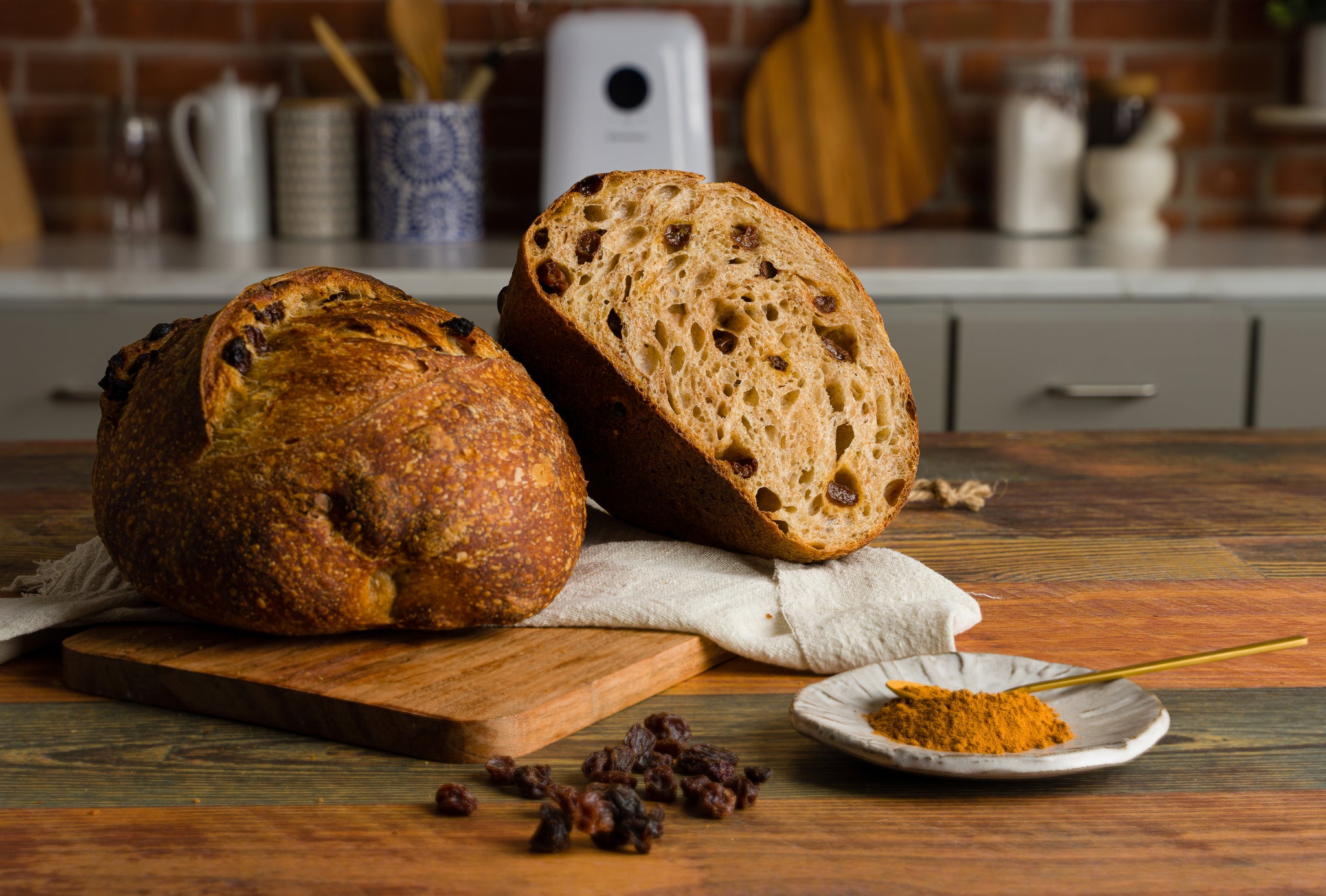 A half loaf of cinnamon raisin sourdough propped up and a whole loaf. A small dish of cinnamon sits to the front right.