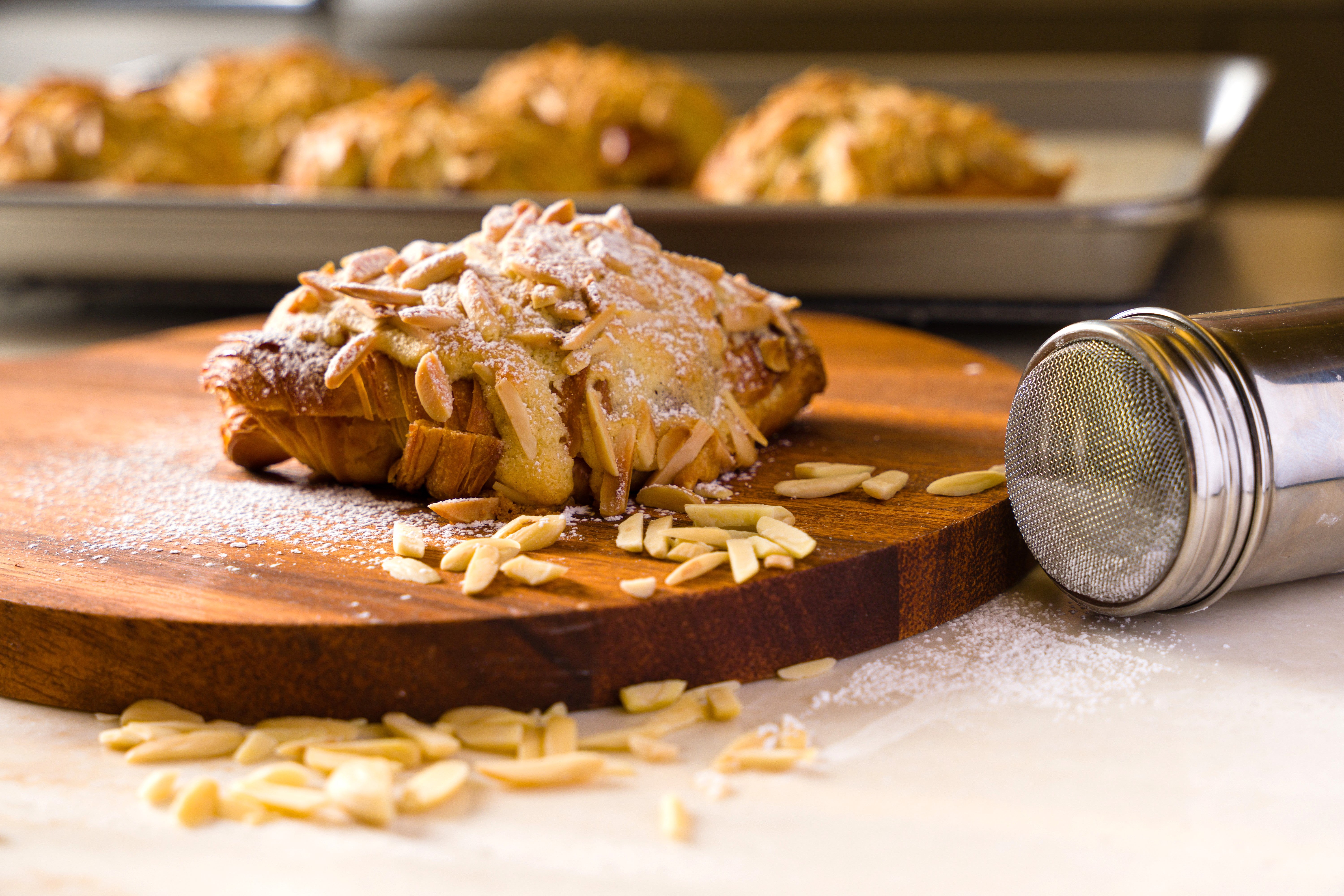 Almond croissant dusted in sugar sits on a round wooden cutting board with a tray of croissants in the background