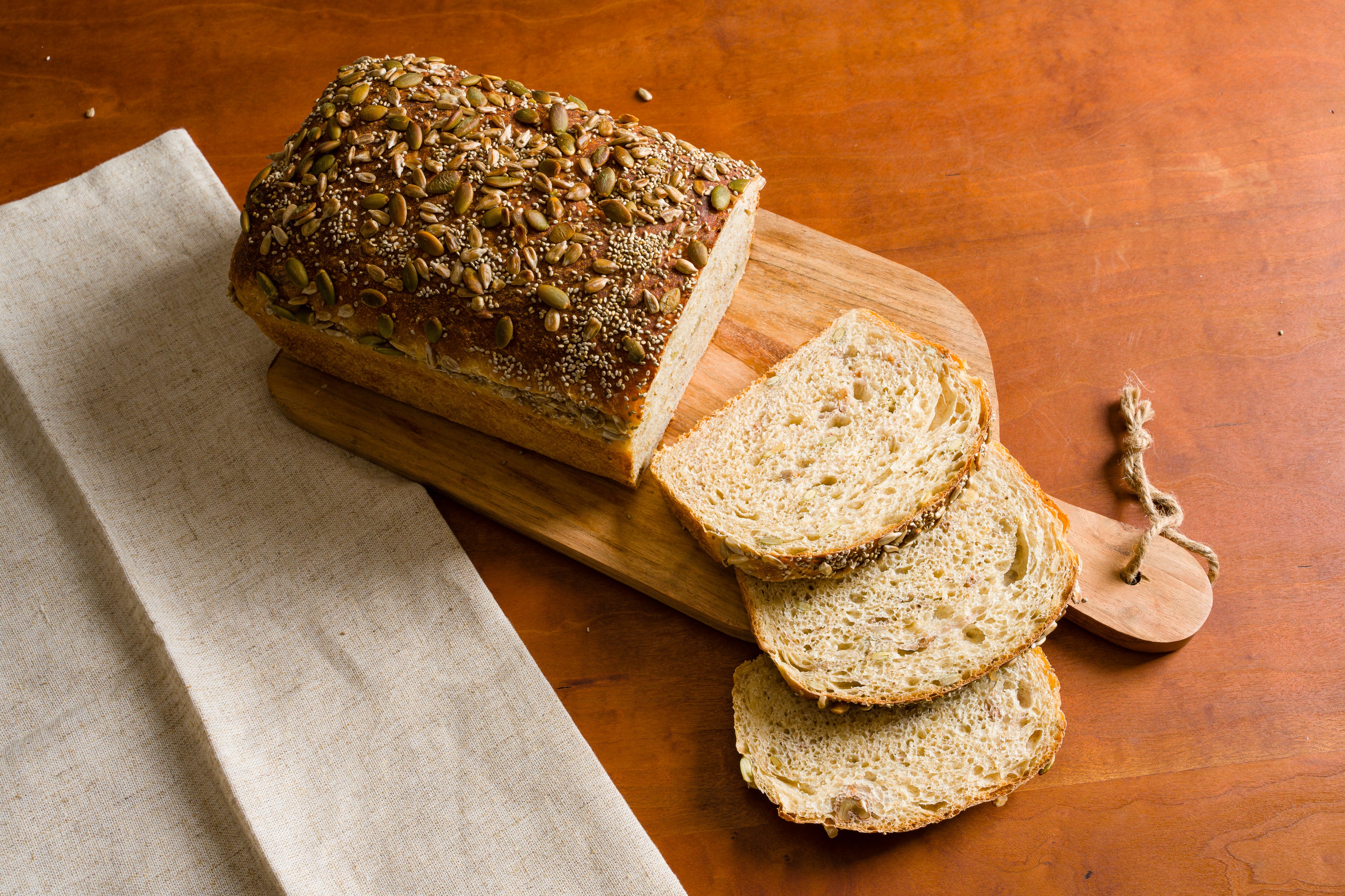 Loaf of sliced buttermilk seed bread on wooden cutting board with beige tea towel to the left