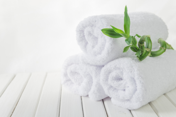 A white wooden floor with 3 white towels rolled up on the right side. A piece of greenery is laid on the top between two towels.