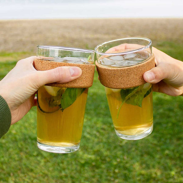 2 people's hands holding glass to go cups filled with clover iced tea, making a cheers gesture.