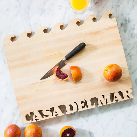 Waves cut out on top of a maple cutting board. The Name Casa Del Mar is cut out along the bottom of the rectangle cutting board. On the board is a knife and some dark red oranges.