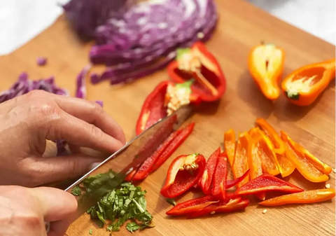 Hands cutting up peppers on wooden cutting board covered in vegatables.