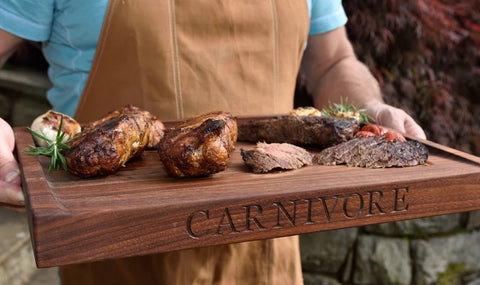 Man holding walnut wood cutting board with different types of meat on it. The word carnivore is engraved on the long side.