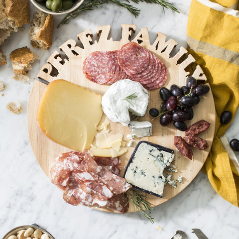 Round Maple Wood Cutting Board with Personalization of Perez Family cut out along the top. Cutting board is on a marble surface with a yellow towel next o it and some pieces of bread on the counter. The board is adorned with slices of salami, a hunk of white cheese, a hunk of bleu cheese, and a wheel of brie with a triangle cut out. Also are some red grapes.