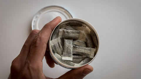 male fingers holding an opened tin of portioned snus with the lid in the background.