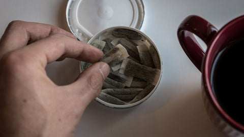 male fingers holding a pouch of portioned snus with a coffee mug in the background.