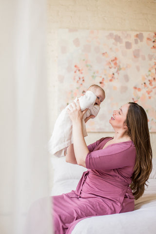 New mom wearing elegant pink hospital gown sitting on bed with newborn held up in air with joy