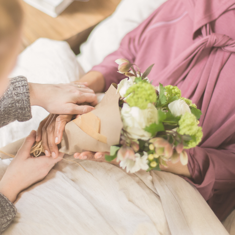 Close up image of woman in pink hospital gown in bed with a second woman's hands handing her a bouquet of flowers