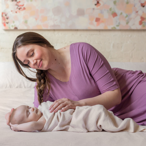 New mama in purple hospital gown with newborn baby swaddled on bed.