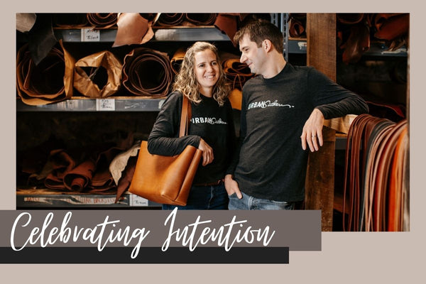 Rachel and Daniel Smucker stand in front of shelves filled with leather. Daniel is looking at Rachel and smiling. White script reads "Celebrating Intention"