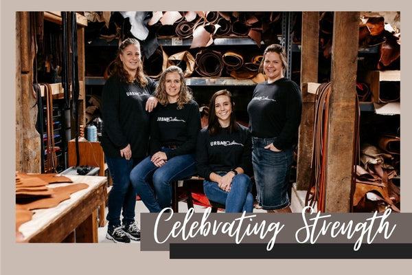 4 women stand together smiling in a shop surrounded by rolls of leather, white script reads "Celebrating Strength"