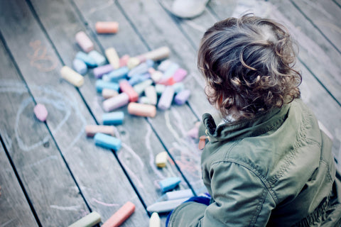 over the shoulder view of a young boy as he looks down on pieces of colorful chalk strewn across a deck