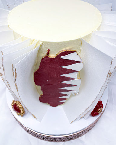 Cream buttercream cake with Qatar Flag Map surrounded by white wafer paper ruffles etched gold with red and gold jewels at the bottom. 
