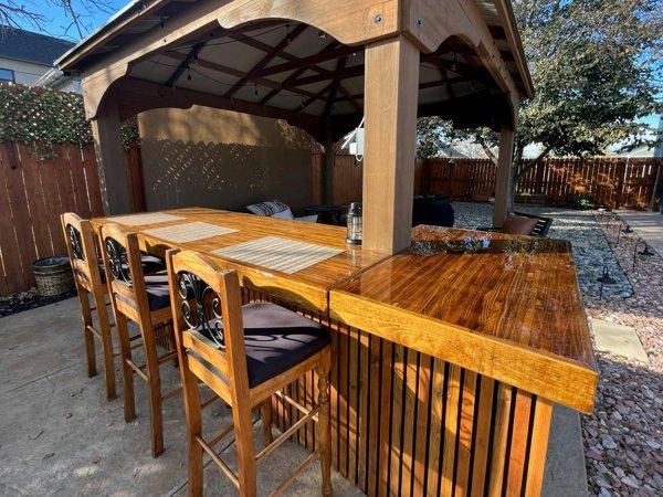 A large outdoor wooden epoxy bar top with a wooden shelter overhead and several wooden chairs.