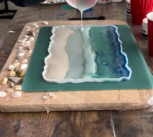 Tinted epoxy resin being poured amidst too other layers of epoxy resin for an epoxy ocean art project.