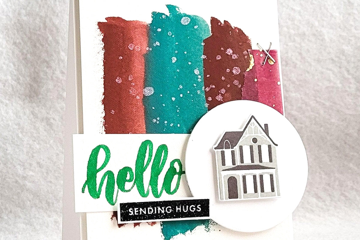A "sending hugs" greeting card with different swatches of deep colors in the background and a die-cut of a two-storey house in the foreground, created with Artistry by Altenew's gouache paint set