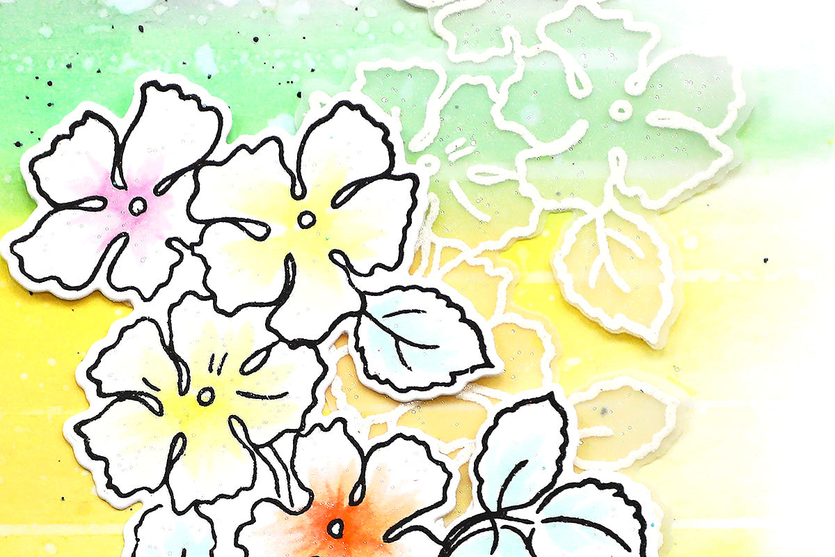 A rainbow ombre background with a faux watercolor effect colored in with the help of ink blending brushes, with a simple outline die-cut of flowers as focal points