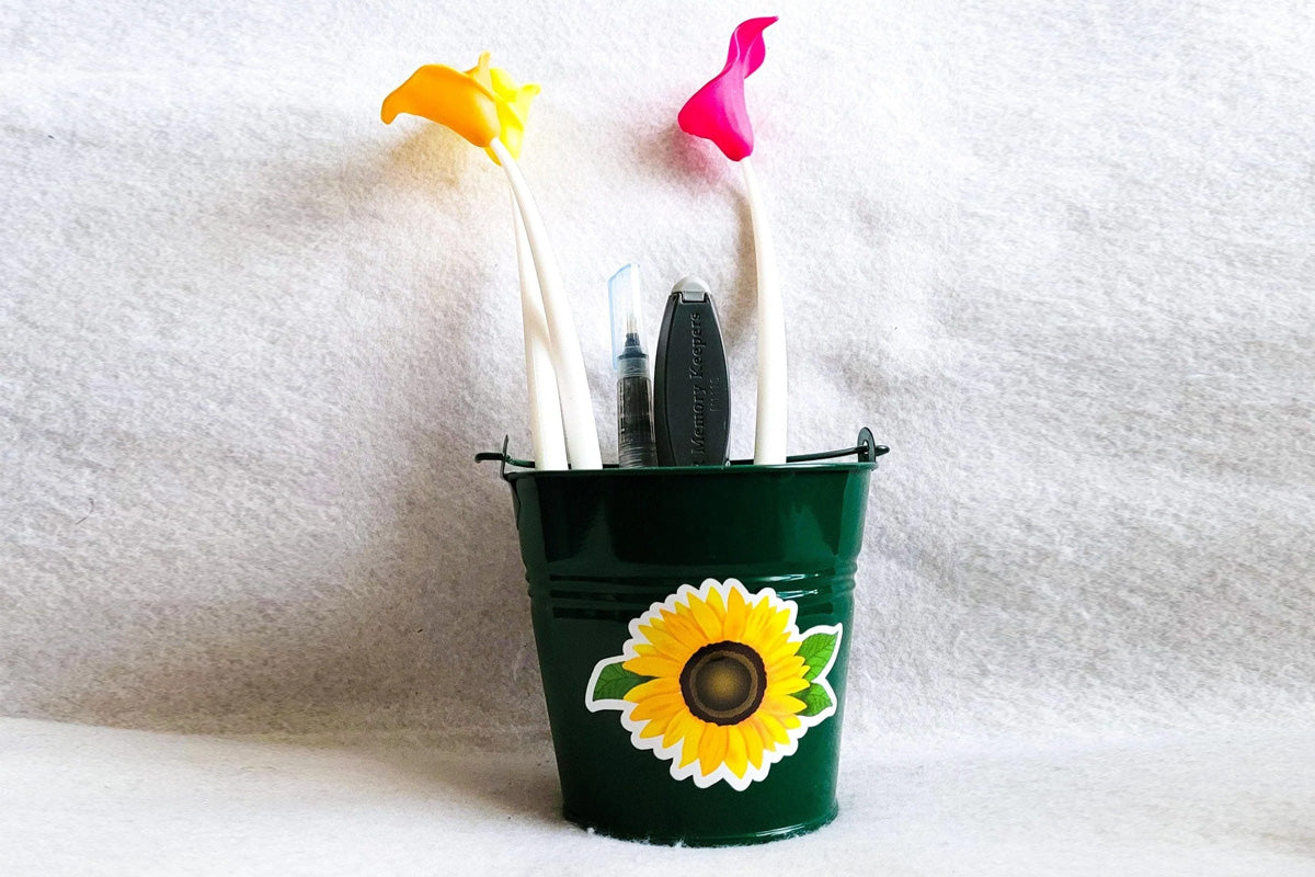 A small green bucket with a sunflower decal, containing pens, markers, and crafting tools