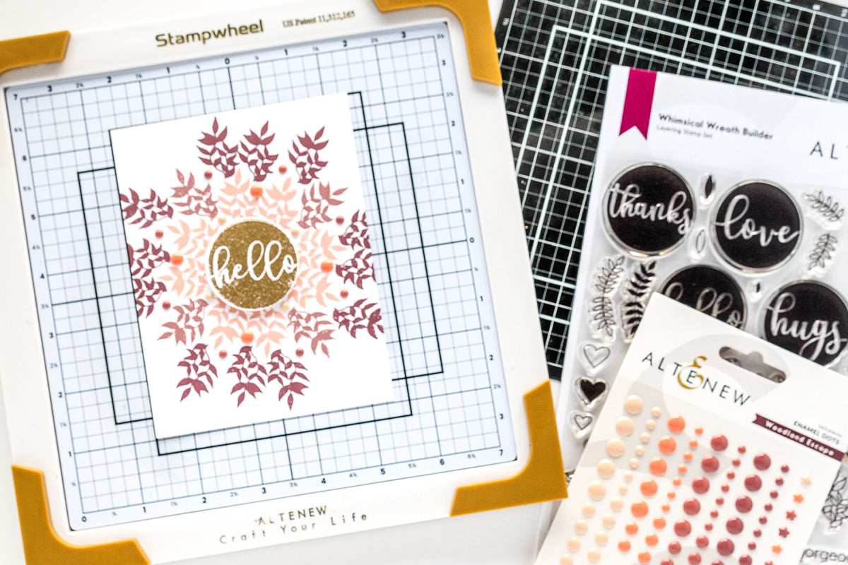 A flat lay photo featuring Altenew's Stampwheel with a wreath-themed greeting card on it, cutting mat, wreath-building stamp set and enamel dots