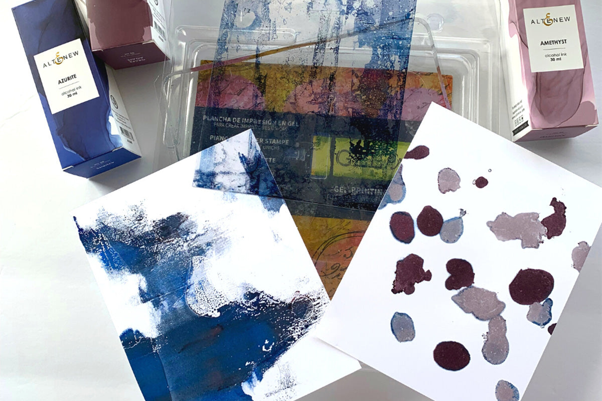 A flat lay photo of 2 alcohol ink panels, a gel plate, and a few Altenew alcohol inks