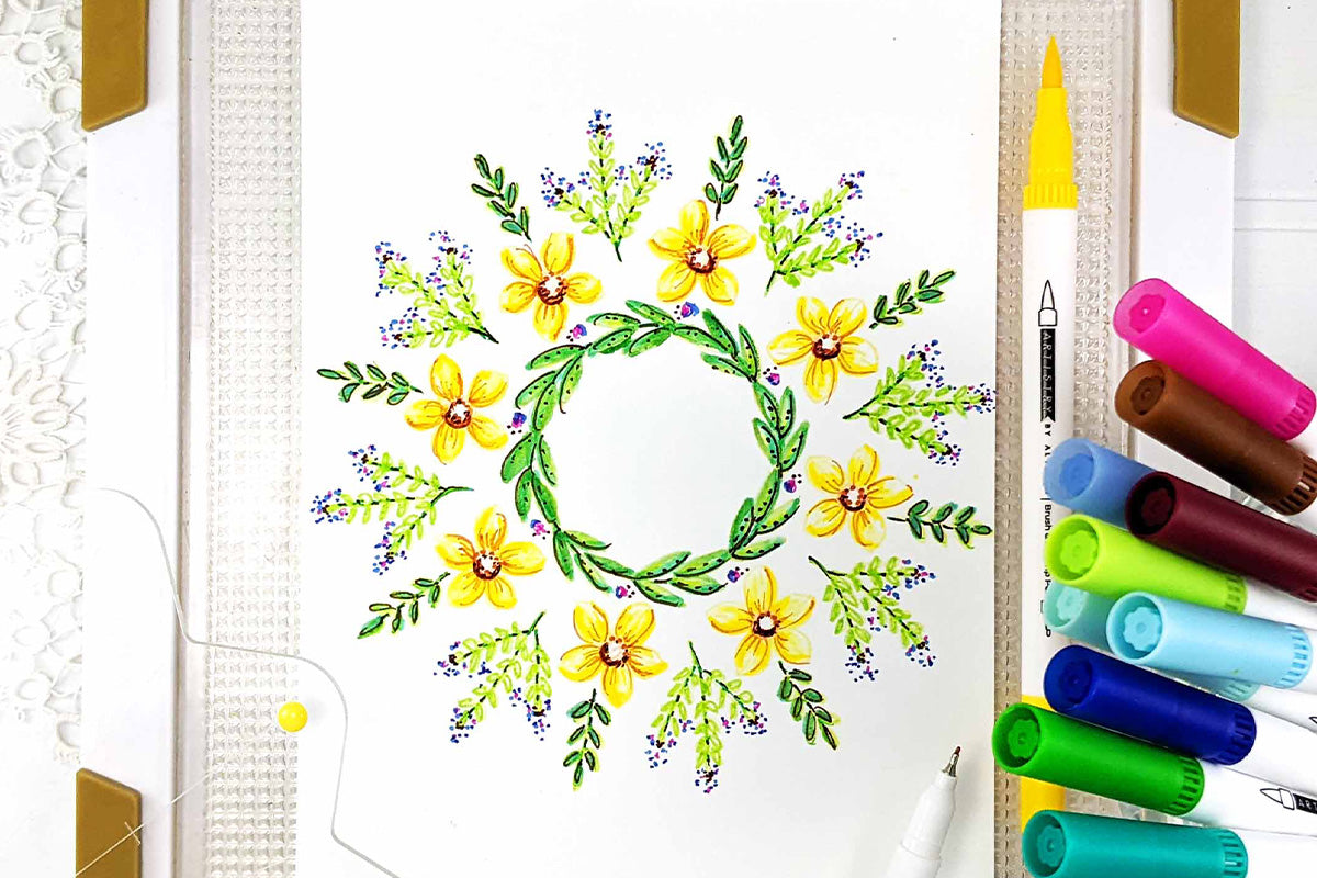 A flat lay photo of a card panel with a floral wreath design colored in with dual-tipped brush pens and placed on top of the Stampwheel