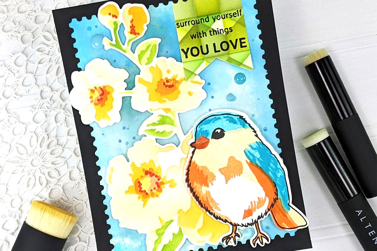 A nature-themed card with a blue background and die-cuts of a colorful bird and a stem of blooming flowers, along with the sentiment "Surround yourself with things you love"