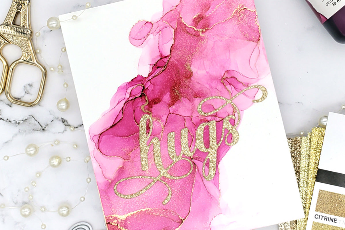 DIY handmade card with an alcohol ink art background and the sentiment "hugs" die-cut in gold glitter paper