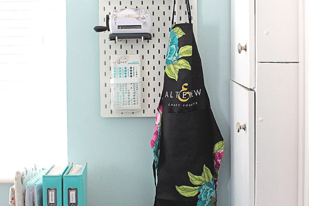 Altenew's Artsy Apron in black with Pink and Aqua flowers, hanging on a peg board in a craft room