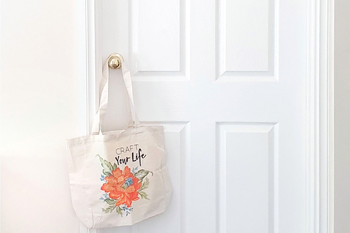 A white door decorated with a craft room decal and Altenew's Craft Your Life tote bag hanging on the door knob