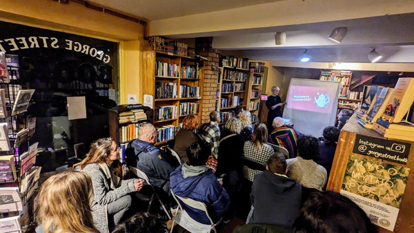 Artist Michelle Collier speaking to a Curiosity Club audience in the bookshop with slides behind about mushrooms