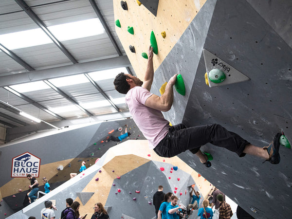 A man in a pink bouldring t shirt is climbing an indoor bouldering wall