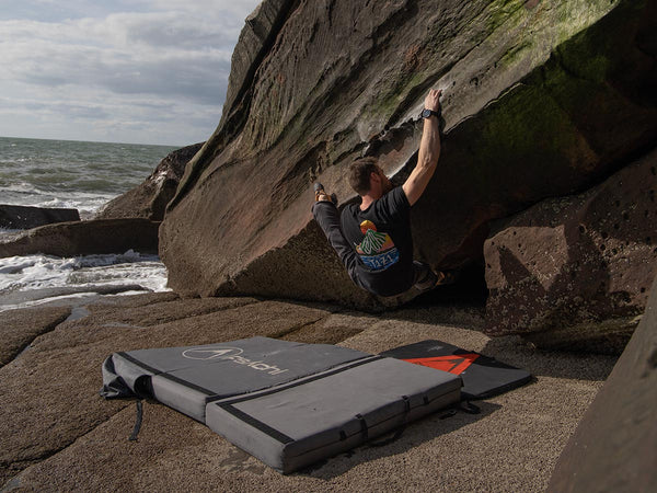 A man in a black boudering t shirt is climbing rock above a bouldering crash pad near the sea