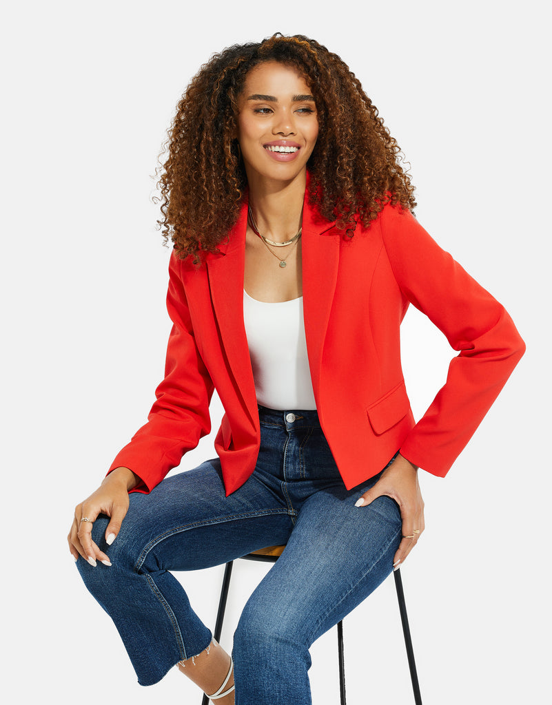 Woman wearing a Threadbare Racing Red Blazer with a white bodysuit and blue jeans, sat on a stool