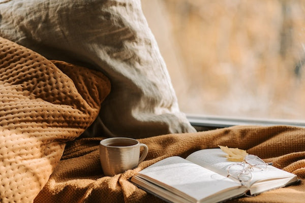 A cozy corner with warm colored throw pillows, blanket, an open book and hot drink