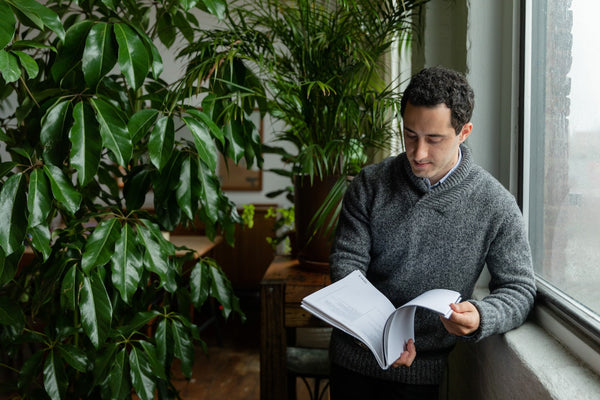 Photo of a Man in a Gray Sweater Reading Near Green Plants