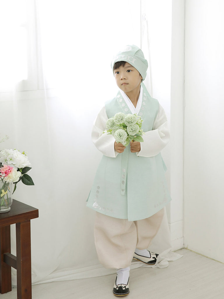 우리의 Soft Cream Boys Hanbok in Mint를 입고 꽃을 들고 비스듬하게 서있는 소년