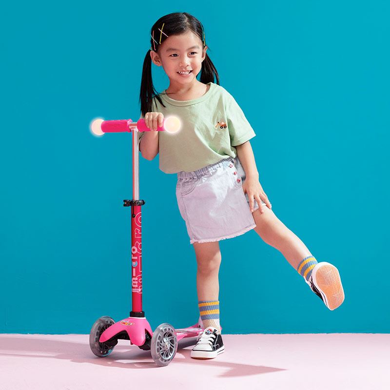 Girl standing beside Mini Micro Deluxe Magic three-wheel kick scooter with LED wheels and light up handlebars