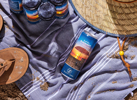 Beach Bum Blueberry sparkling wine can on a beach towel with sun hat, flip flops, and six pack of canned wine.