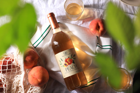 A bottle of Tanglewood Winery's Peach wine lying on the picnic blanket with a bag of fresh peaches and two glasses of wine.