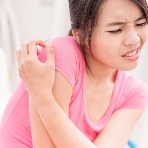 A women in a pink shirt scratching the itchy skin on her shoulder.