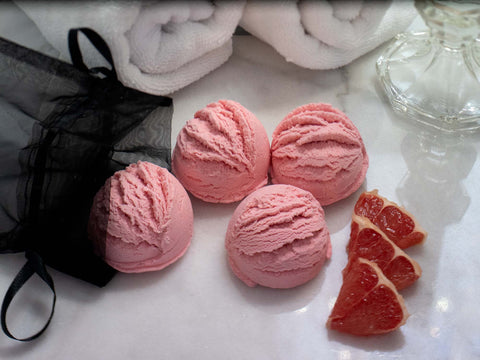 Pink solid botanical bubble bath in the shape ice cream scoops with a black organza bag, white rolled up towels and slices of pink grapefruit.