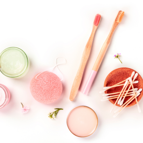 Pink bristle bamboo toothbrushes, wooden Q-tips in and bowl and pink konjac sponge on a white background.
