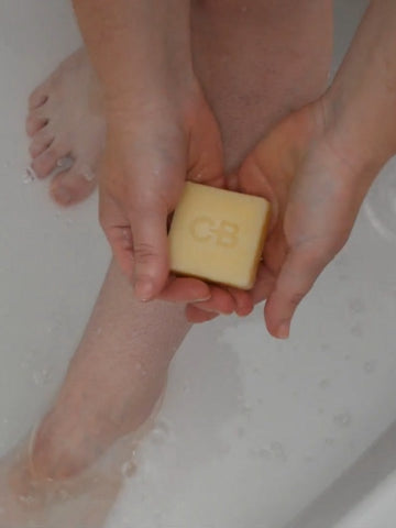 Holding a bar of Caley-Beth Shave balm with two hands, before applying it to the lower leg in the bathtub.