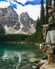 Man and dog standing by lake with mountains in the background