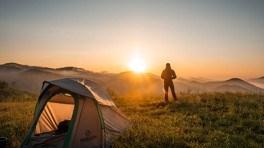 Person overlooking a mountain top with the sun setting and their tent set up for camping