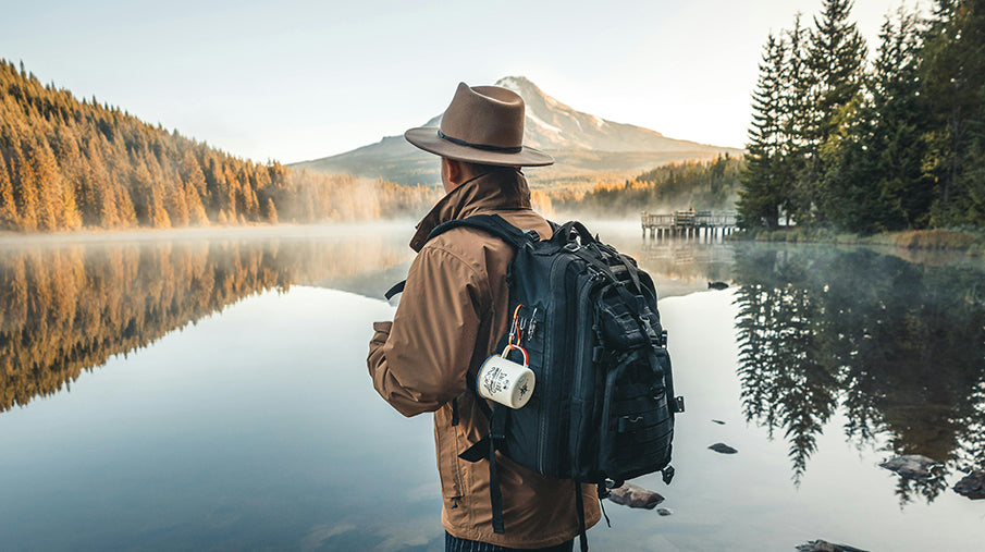 Man with backpack and hat looking out over a lake surrounded by mountains